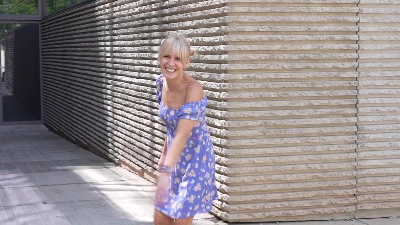 Uma mulher com um vestido floral azul está dançando em um cenário urbano ensolarado perto de uma parede texturizada.