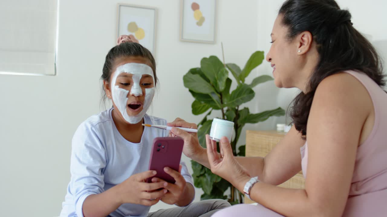 Mamma afroamericana e figlia adolescente che creano un legame sul letto, la mamma intinge il pennello nel barattolo applicando una maschera