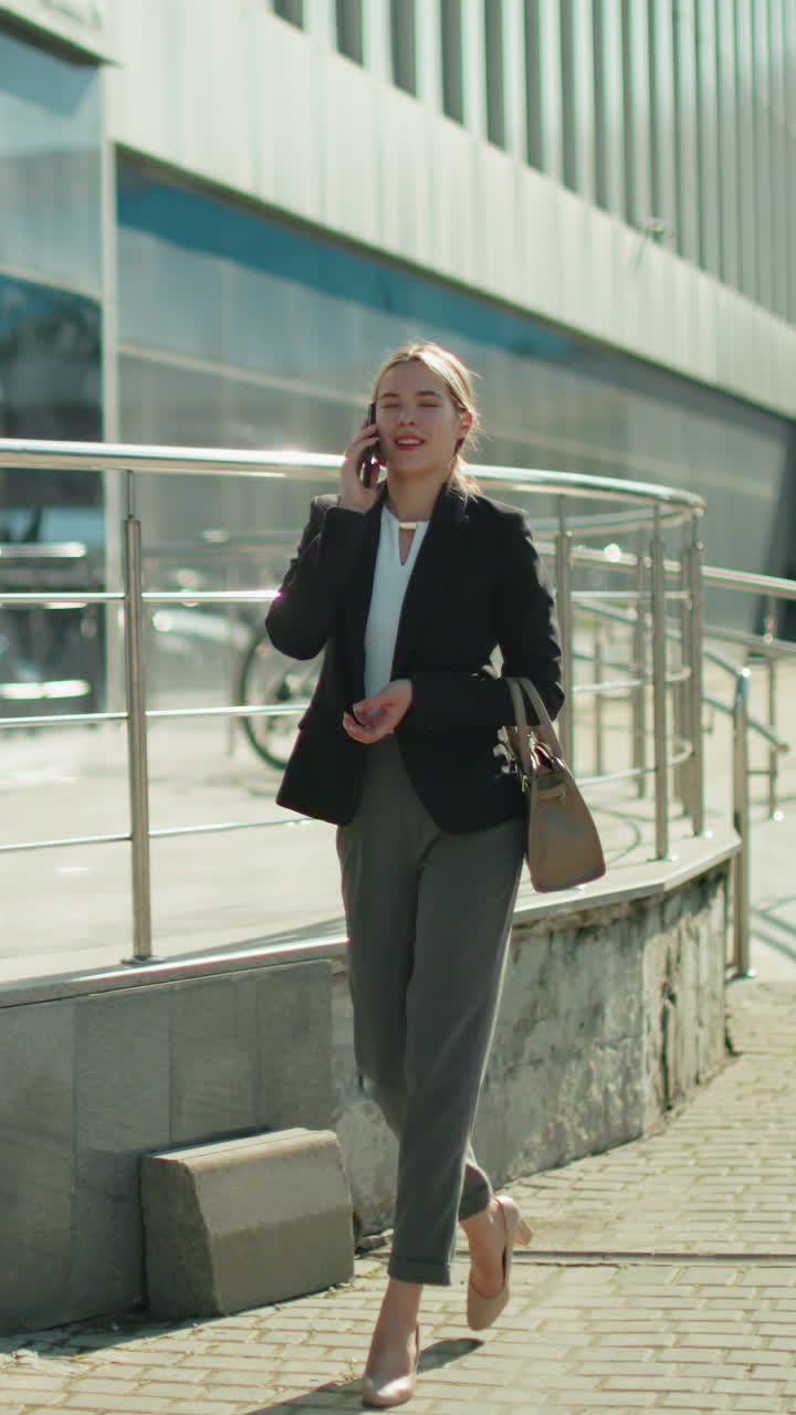 Professional business woman walks home while talking on phone, dressed in formal outfit, holding handbag, with urban glass building reflecting surroundings