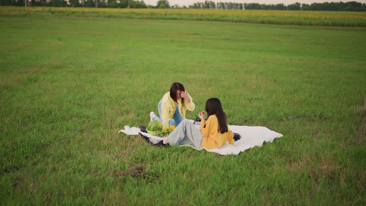 Women sharing peaceful moment, Calm women converse in nature, Pair of women chatting softly on blanket in scenic field, Two friends relaxed on blanket amidst wide open meadow under cloudy sky