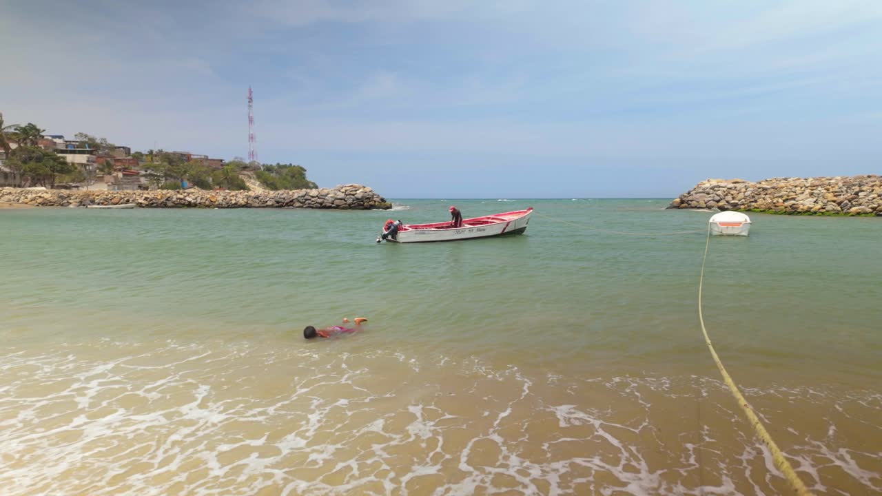 A child swims in the sea and walks toward the shore, with a fishing boat in the background.