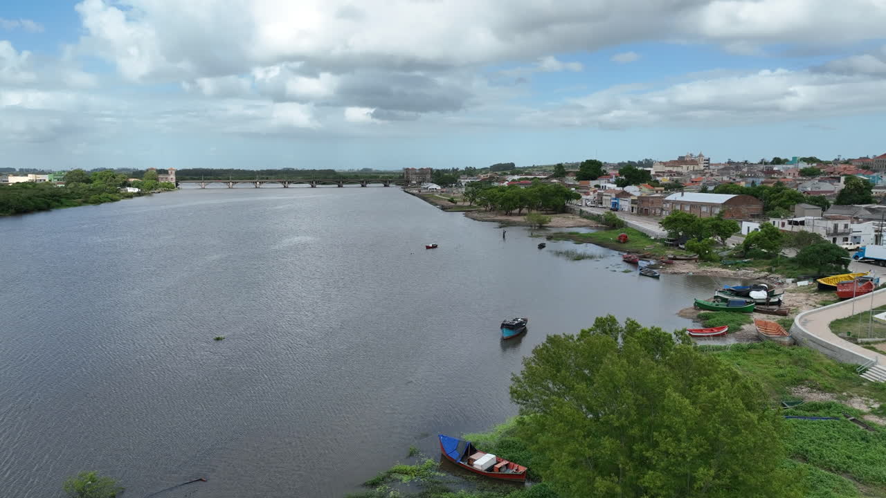 Volo cinematografico a bassa quota con drone sopra le tradizionali barche da pesca sulla riva del fiume Jaguarão. Inquadra lo storico ponte Mauá e il confine con l'Uruguay sullo sfondo. Master professionale 4K a 60 fps