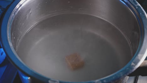 mano agregando un cubo de especias marrones al agua caliente en una olla gris en la estufa