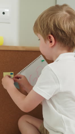 little boy pins note to cork board sitting on table with bare legs slow motion. blond preschooler plays with stationery. educational process at home side view