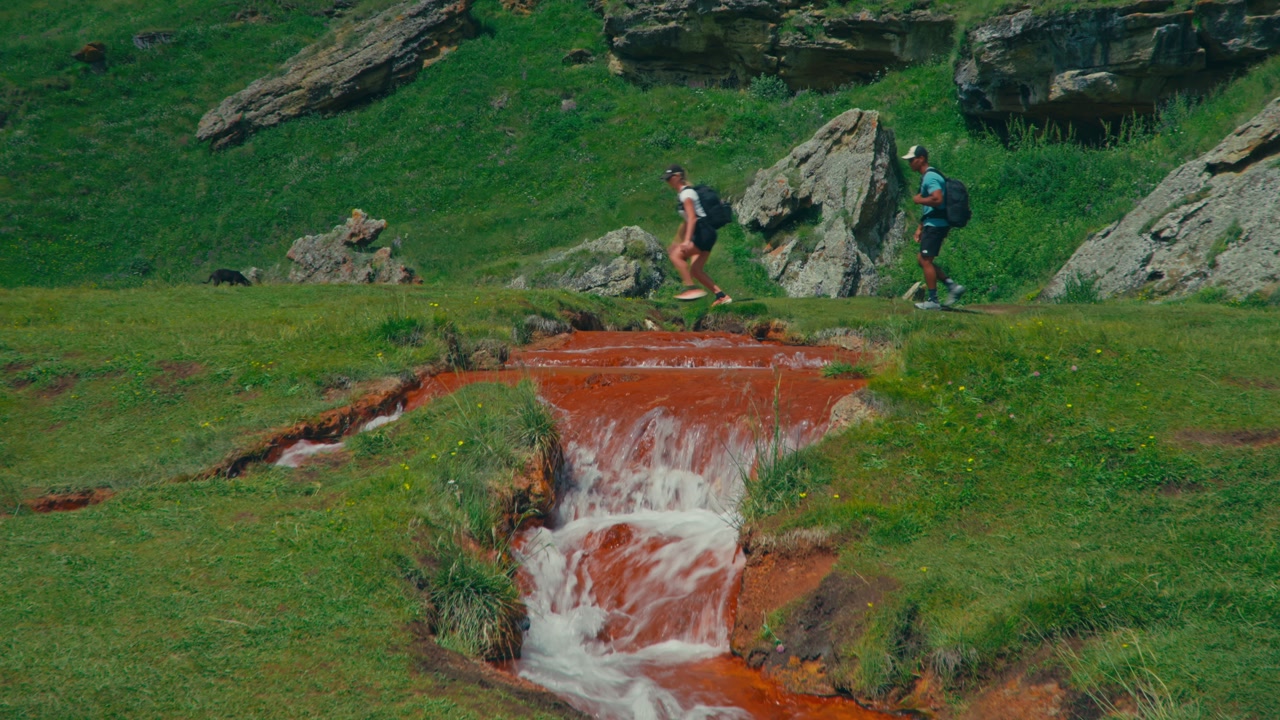 Two hikers jump over a colorful red mineral spring with a small waterfall in a lush mountainous landscape in Truso Valley, Georgia, capturing adventure and unique geology.