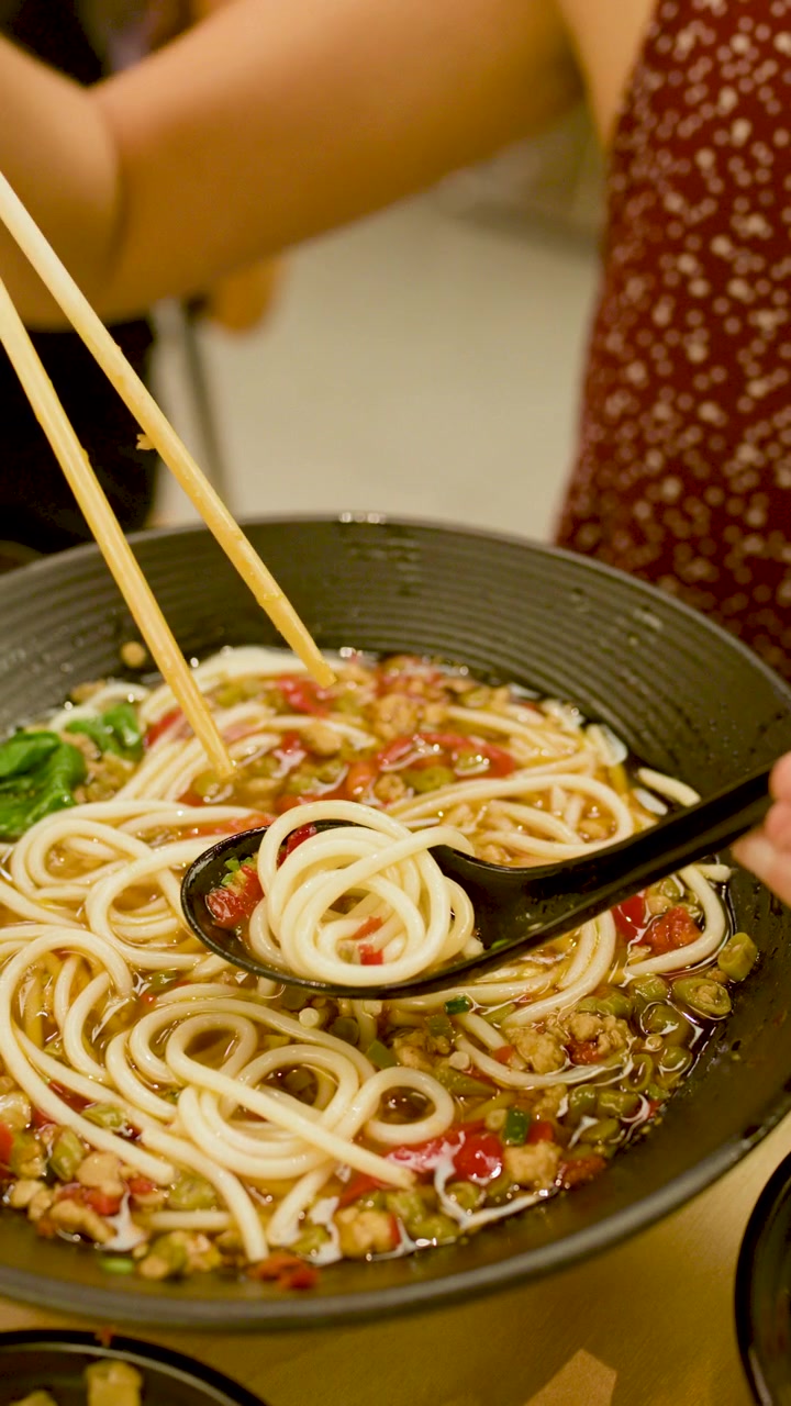Una persona usa le bacchette per sollevare una zuppa di noodle piccante da una ciotola nera sotto una calda illuminazione interna.