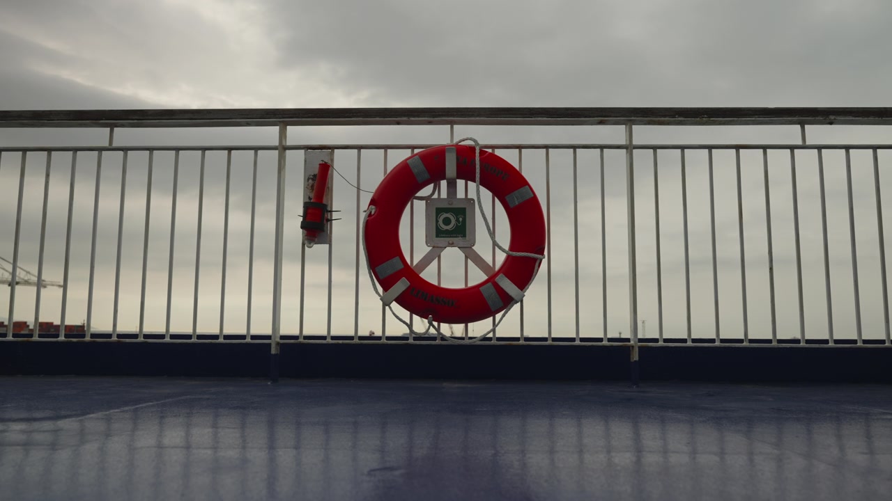 Red life preserver ring fixed to metal railing on ferry deck with grey overcast sky and port behind