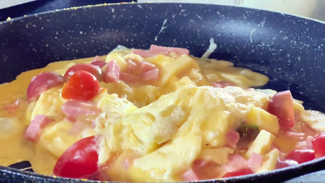 Primo piano di una spatola rossa che mescola una frittata con prosciutto a cubetti e pomodori in una padella. Ideale per ricette per la colazione, pubblicità di pentole e video culinari