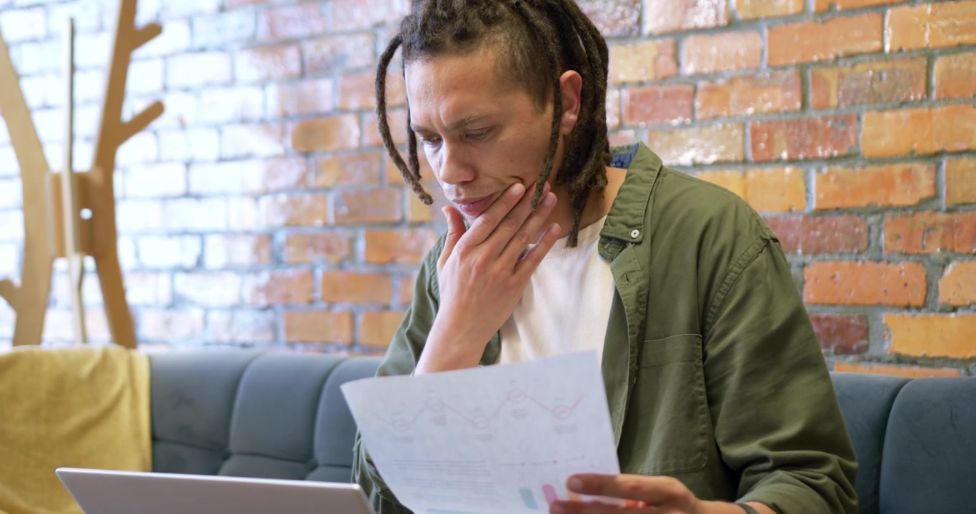 Uomo afroamericano che tiene grafici stampati e li confronta con un laptop sul divano mentre analizza i dati.