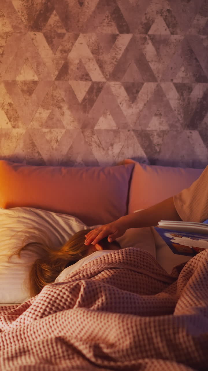 Long haired woman with book strokes head of little sleeping child covered with soft duvet near toy playhouse with glowing lamp in bedroom slow motion