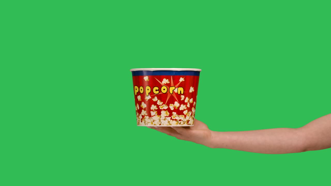 Female hands giving a bucket of popcorn close up. Woman points to the box and makes a thumbs up gesture. A container with a delicious snack on the background of a green screen chroma key. Slow motion