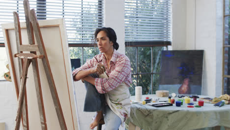 biracial female artist in apron holding brush and looking at canvas in studio, slow motion