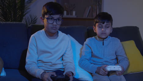 two young boys sitting on sofa at home playing with computer games console on tv holding controllers late at night 2