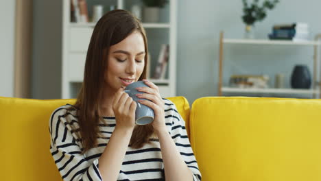 retrato de la joven mujer guapa tomando café o té y sonriendo mientras se sienta en el sofá en casa