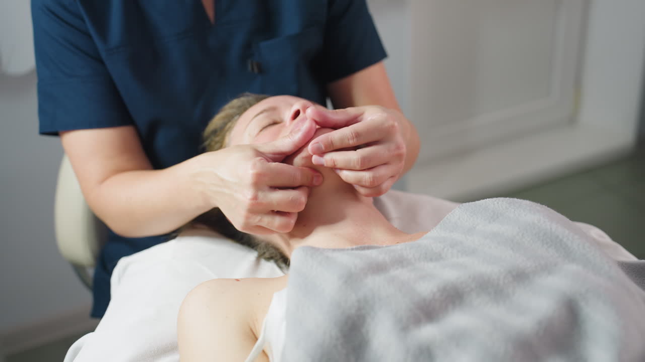Participant lying on massage bed face up as skin care expert cradles jawline gently with fingertip support under soft lighting for relaxation comfort and skin rejuvenation during facial therapy