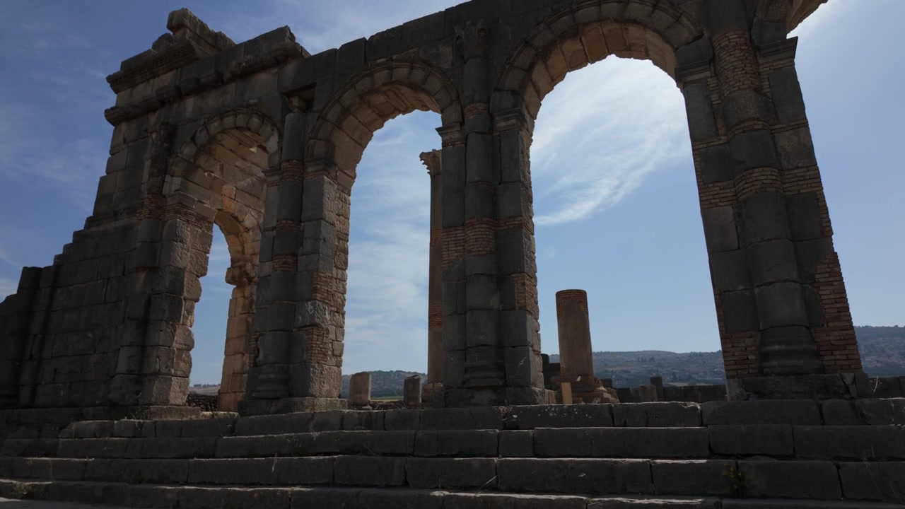 Maanpintatason elokuvamainen näkymä Volubilisin basilikan monumentaalisista kaarista, korostaen roomalaista oikeudellista arkkitehtuuria Marokon UNESCOn maailmanperintökohteessa.