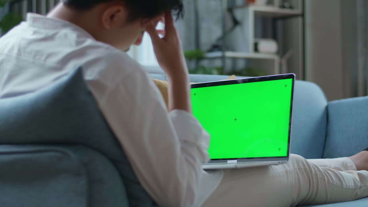 hombre cansado mientras yace en el sofá y usa una computadora portátil de pantalla verde en la sala de estar