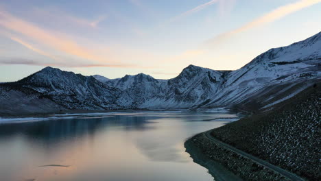 Vibrant-evening-aerial-shot-over-lake-water-and-mountain-range-covered-in-snow