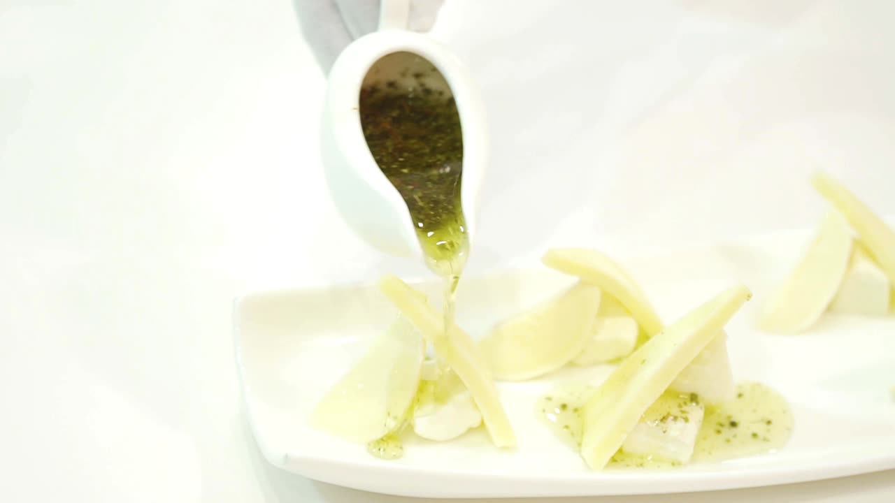 Hand with the white cup with green sauce pouring out on the pieces of cheese nicely decorated on a plate. Delicious appetizer for guests in the restaurant. Close-up