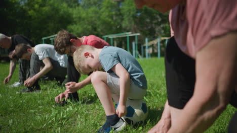 close-up of a family kneeling on a grassy field, with the youngest child sitting on a soccer ball while others focus on tying their shoelaces