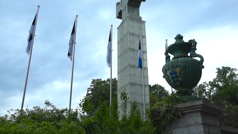 gorgeous panning up footage of tallinn freedom square cross of liberty statue or monument in central city during a cloudy day with estonian flags around and large sculptures with estonian coat of arms