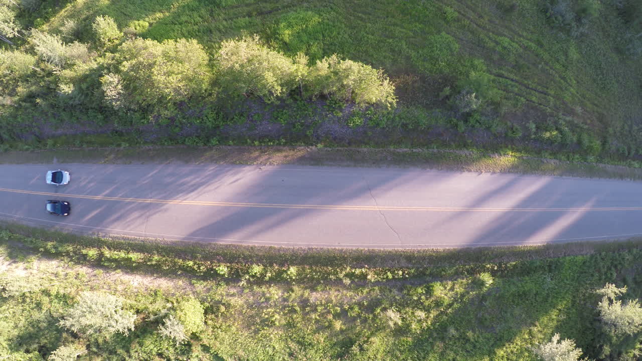 Sports cars cruise down a country road in the remote wilderness, from the perspective of a drone overhead at sunset.