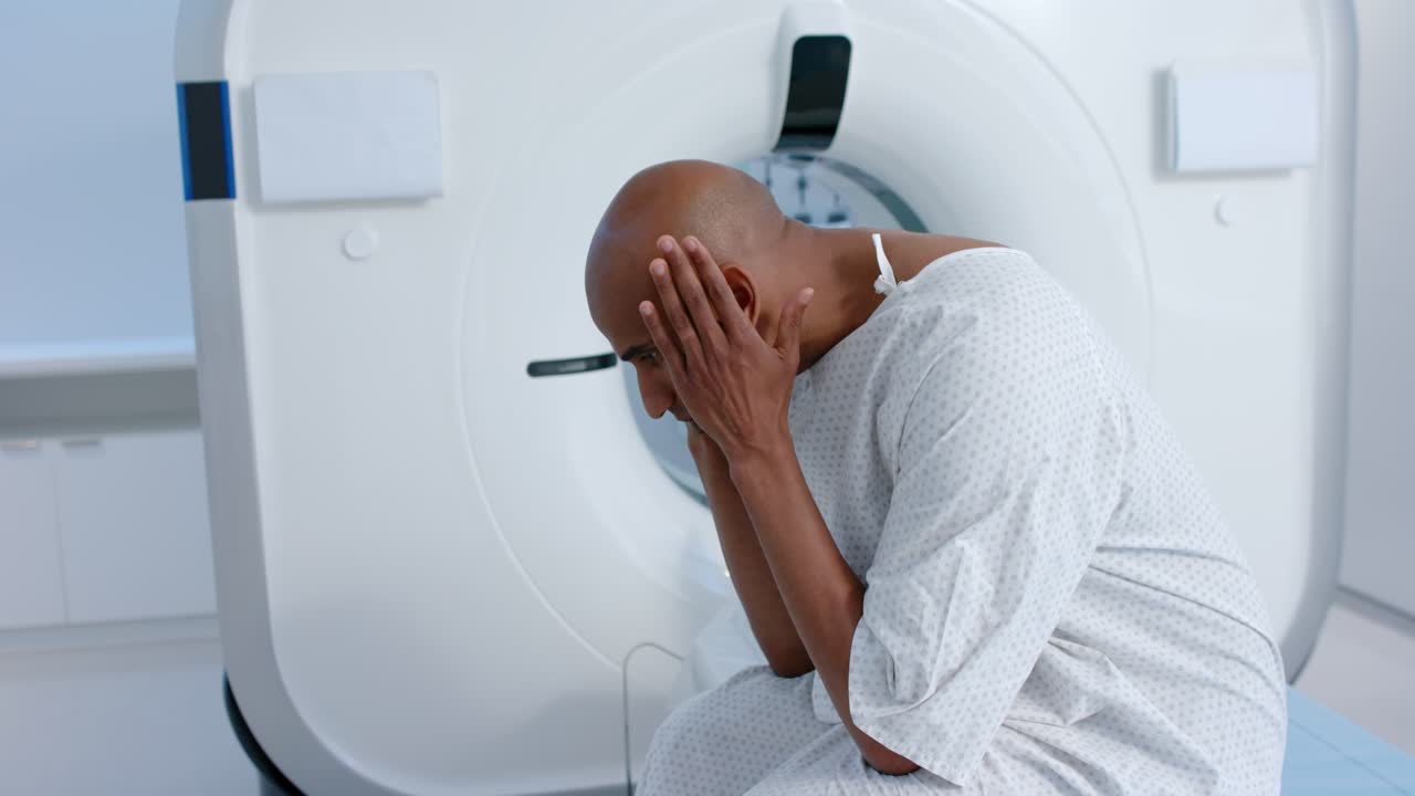 Uomo afroamericano in camice seduto al tavolo da visita accanto a uno scanner, con il volto tra le mani, preparandosi per la scansione