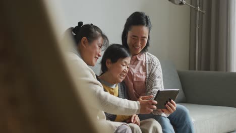 smiling senior asian woman using tablet with adult daughter and granddaughter embracing