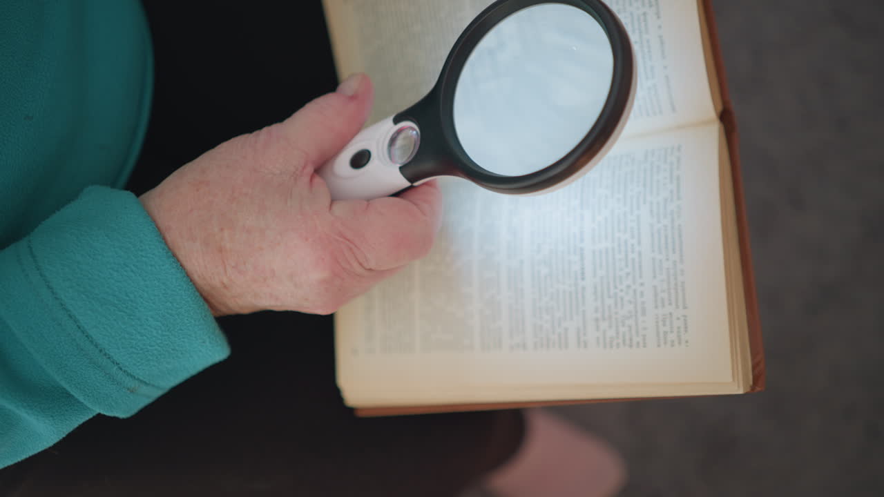 Senior woman using magnifying glass to read book, close-up of hand holding magnifier over text, highlighting focus on reading, with natural light illuminating book and hands