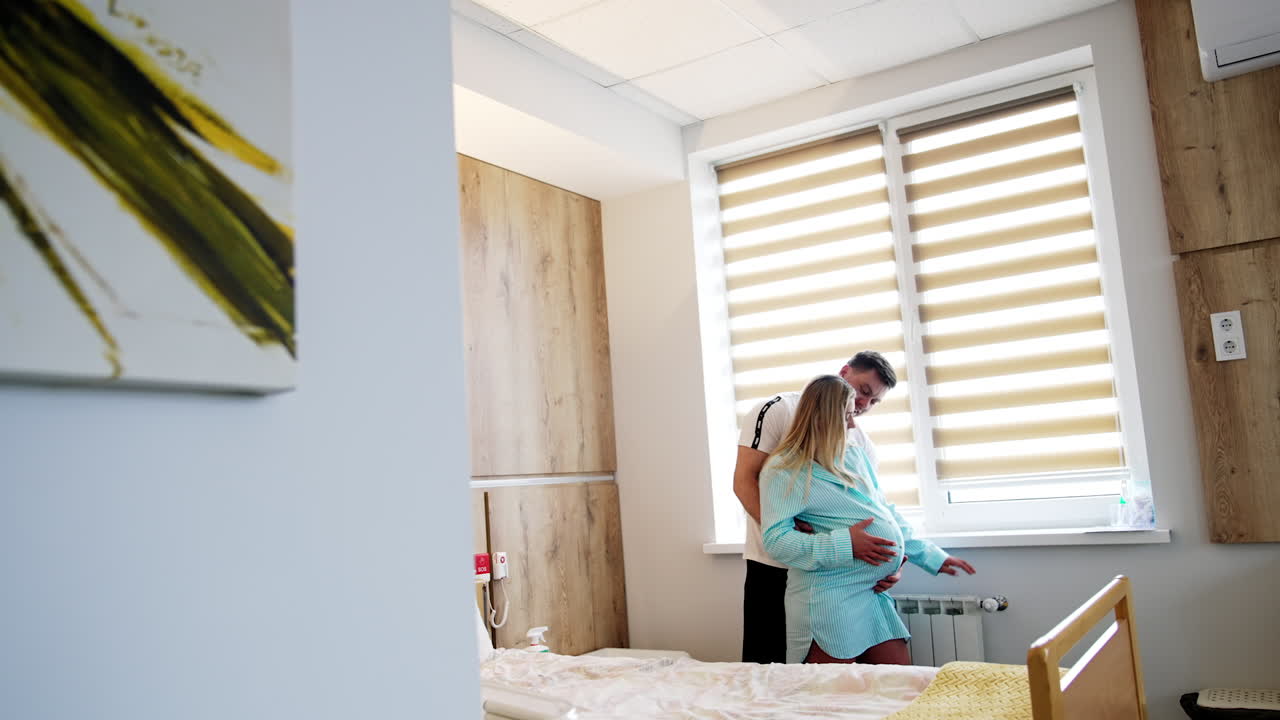 Comfortable cozy ward in the maternity hospital. Loving couple expecting a baby stand near the window.