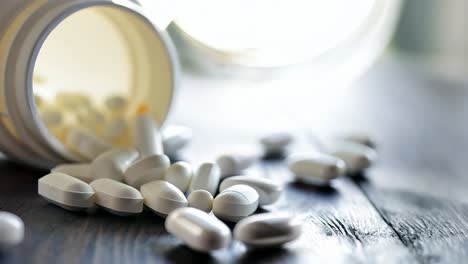 white pills are spilling from a white plastic container lying on its side on a dark wooden table, with a bright light source in the background