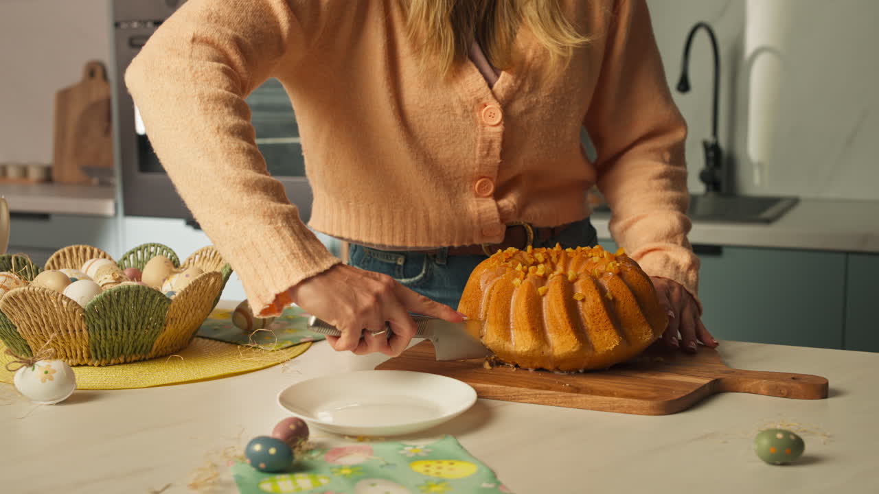 Woman Slicing Golden Bundt Cake On Wooden Board In The Kitchen, With Painted Easter Eggs Scattered On The Counter. medium shot
