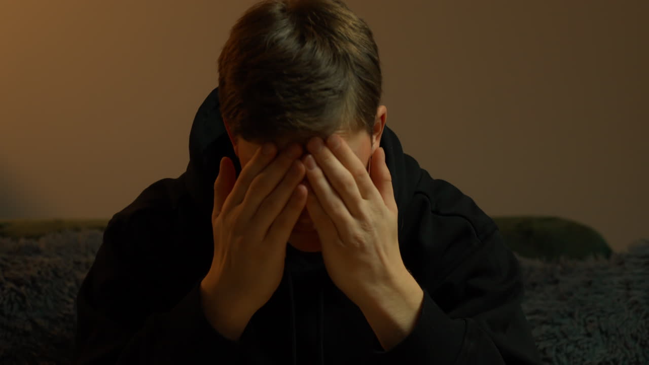 Young man covering his face with his hands, expressing deep despair, loneliness, crying, mental health issues, and emotional pain in a dimly lit indoor setting, slow motion shot
