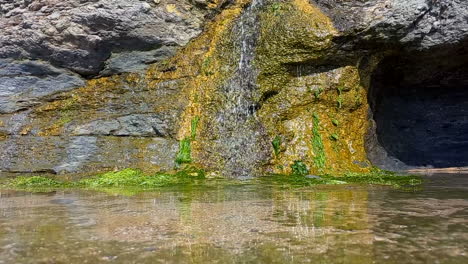 Water-running-down-the-rocks-in-sea-caves-near-the-coast,-forming-a-mirror-of-water-and-green-slime-on-the-rocks