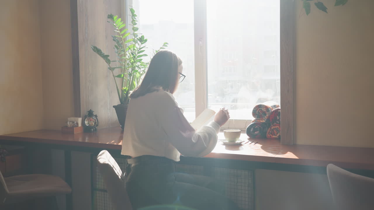 Rear view of woman studying near bright window with open book and steaming mug on table, natural sunlight casting soft ambient glow through interior filled with plant