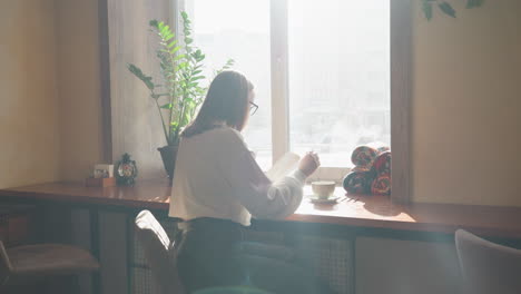 rear view of woman studying near bright window with open book and steaming mug on table, natural sunlight casting soft ambient glow through interior filled with plant