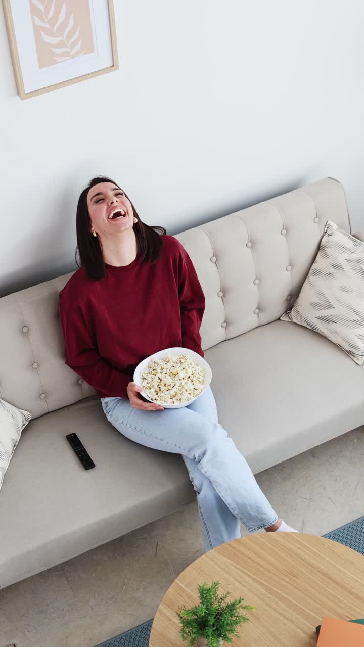 Glad ung kvinde griner, mens hun ser tv og spiser popcorn