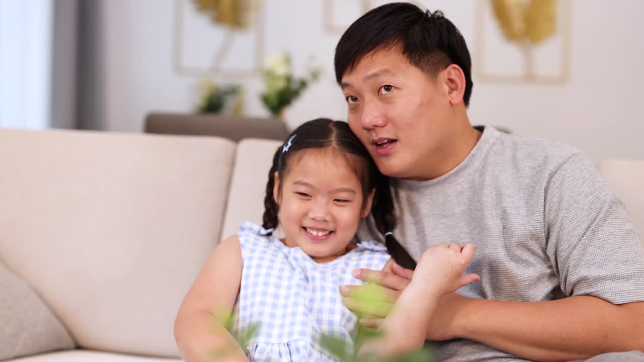 Asian father and daughter share a loving, protective moment on sofa watching movie at home