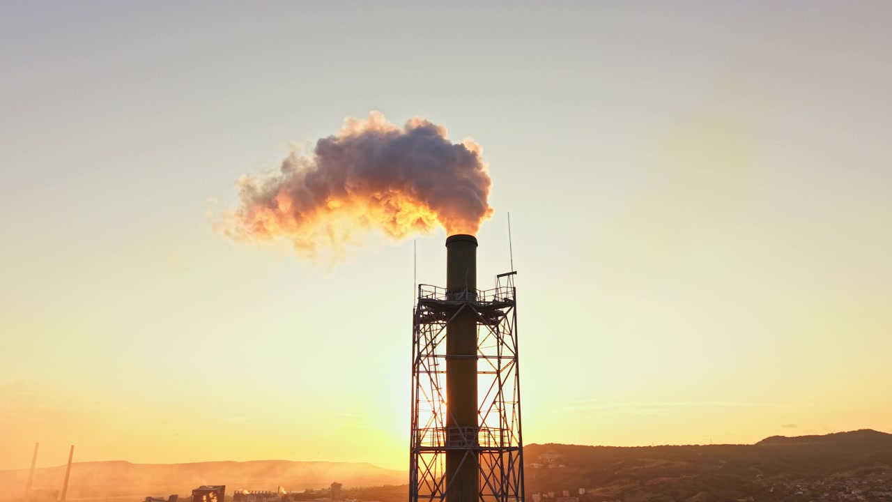 Il fumo sale da una ciminiera di fabbrica al tramonto su un paesaggio urbano con colline sullo sfondo