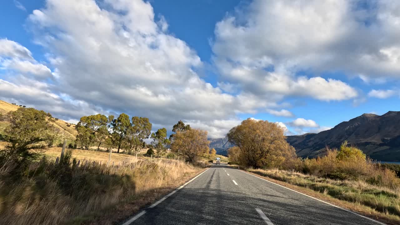 Car travels along rural road with autumn trees, mountains, and dynamic clouds under natural daylight