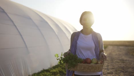 Blonde-woman-walking-near-the-greenhouse,-carrying-fresh-vegetables-or-plants-outdoors