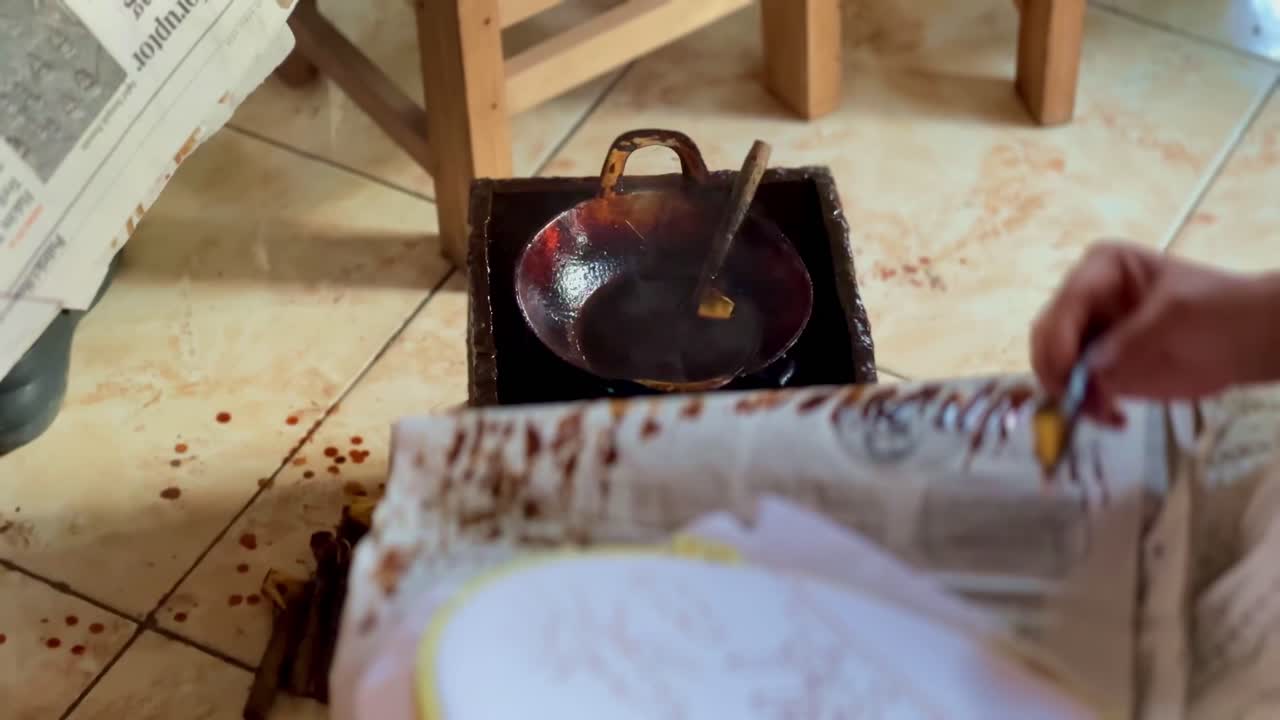The hand is holding a tool "canting" for making batik which is placed in a small pan filled with liquid wax