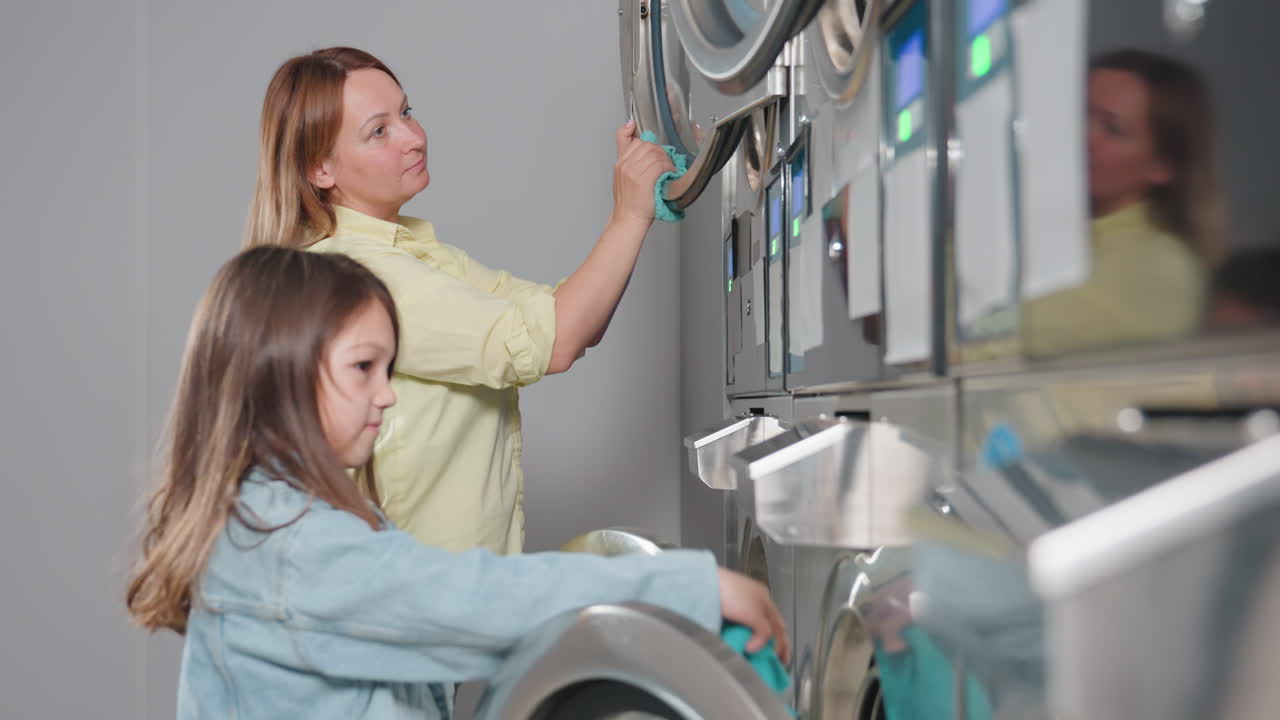Side view laundry owner guides daughter cleaning stainless dryers, showing wipe turquoise cloth, teamwork lesson inside laundromat, hygiene focus, supervision, row of machines with glowing screens