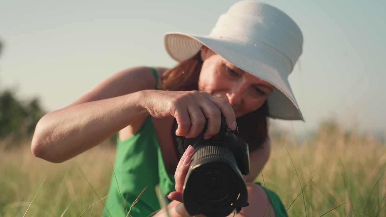 close up of woman in green dress holding camera kneeling in tall grass as she takes picture of wild plant in golden hour light with warm focus and gentle concentration on subject