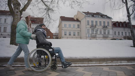 Side-view-of-happy-Caucasian-woman-taking-her-disabled-friend-in-wheelchair-for-a-walk-in-the-winter-city-and-talking-together
