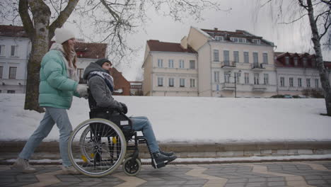 side view of happy caucasian woman taking her disabled friend in wheelchair for a walk in the winter city and talking together