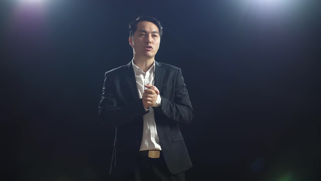 Asian Speaker Man In Business Suit Holding His Hands Together And Showing Index Fingers Up While Speaking In The Black Screen Studio