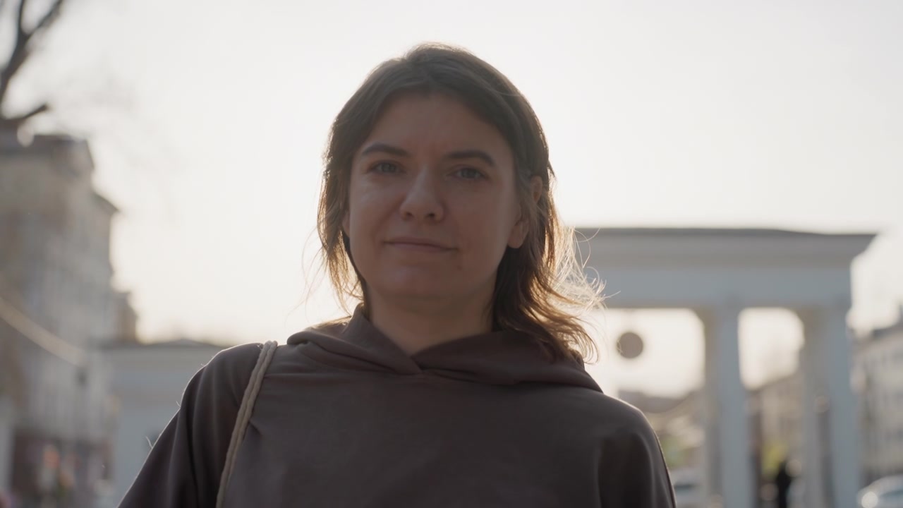 Donna in piedi all'aperto in una piazza pubblica, che indossa una felpa con cappuccio scura e porta una borsa a tracolla. Lo sfondo presenta architettura urbana con edifici e un arco, sotto una luce diurna soffusa.