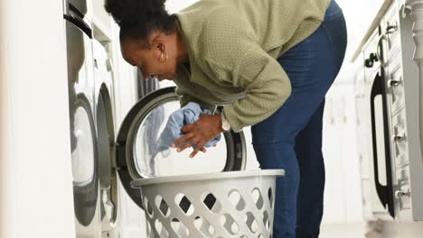 happy african american senior woman putting laundry in washing machine in sunny kitchen, slow motion