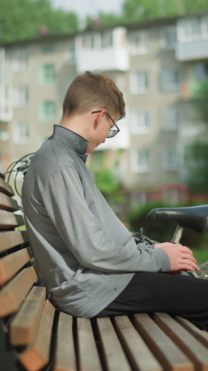 회색 과 검은 바지를 입은 소년은 자전거를 주차하고, 평화로운 공원에서 나무 벤치에 앉아, 뒤로 기대어, 다리를 고, 자전거에 눈을 돌리고, 초록색으로 둘러싸여 있습니다.